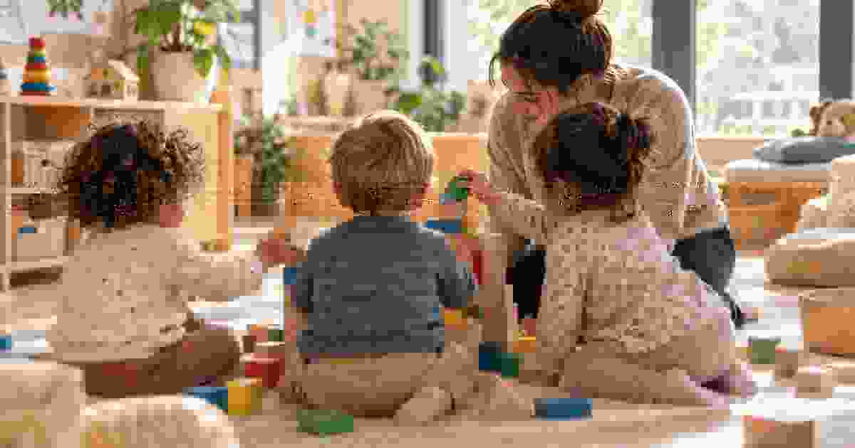 Drei kleine Kinder bauen gemeinsam mit Begleitung einer pädagogischen Fachkraft einen Turm aus Bausteinen